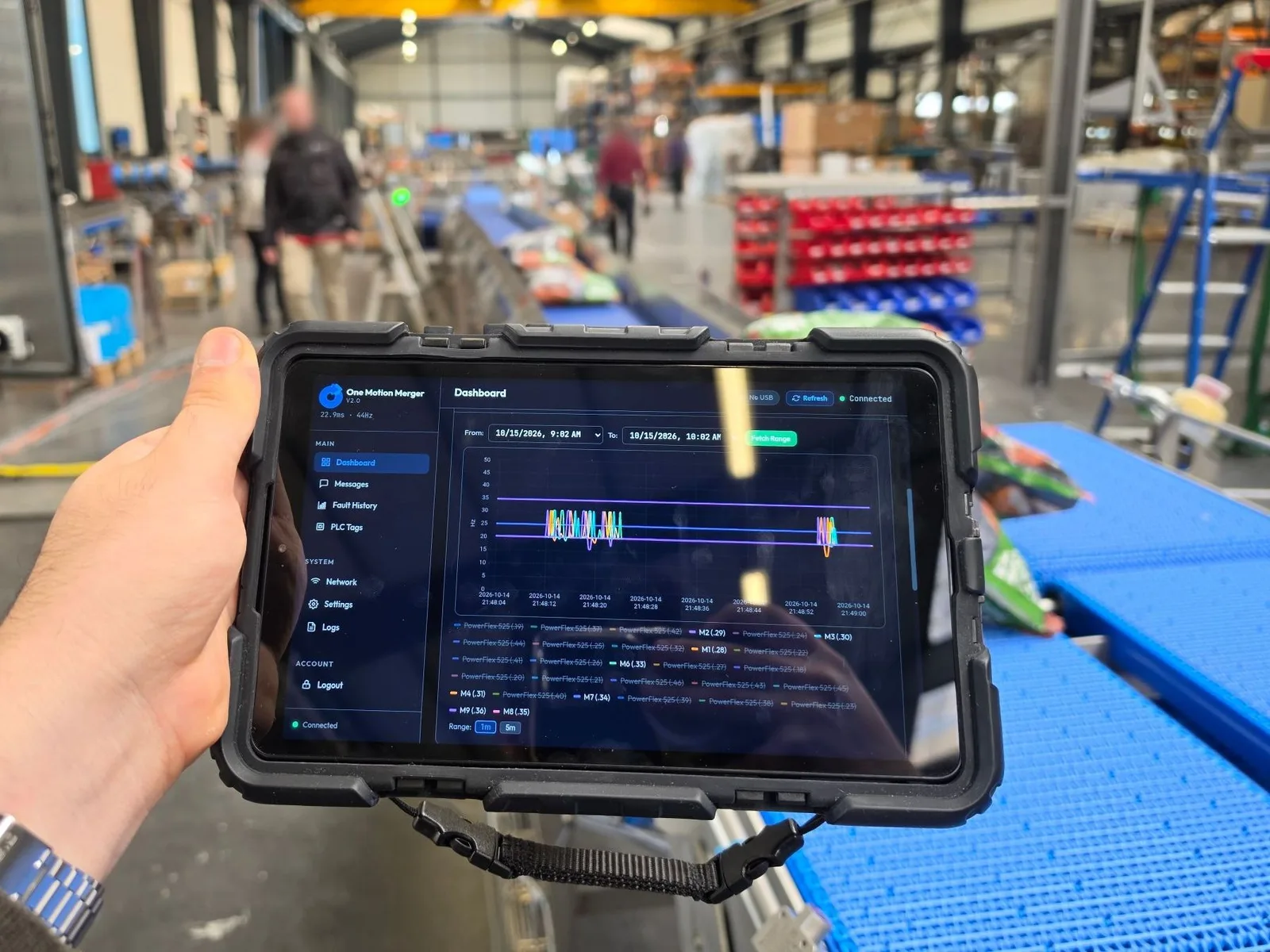 CorTex dashboard on a rugged tablet held beside a running blue modular-belt conveyor in a plant